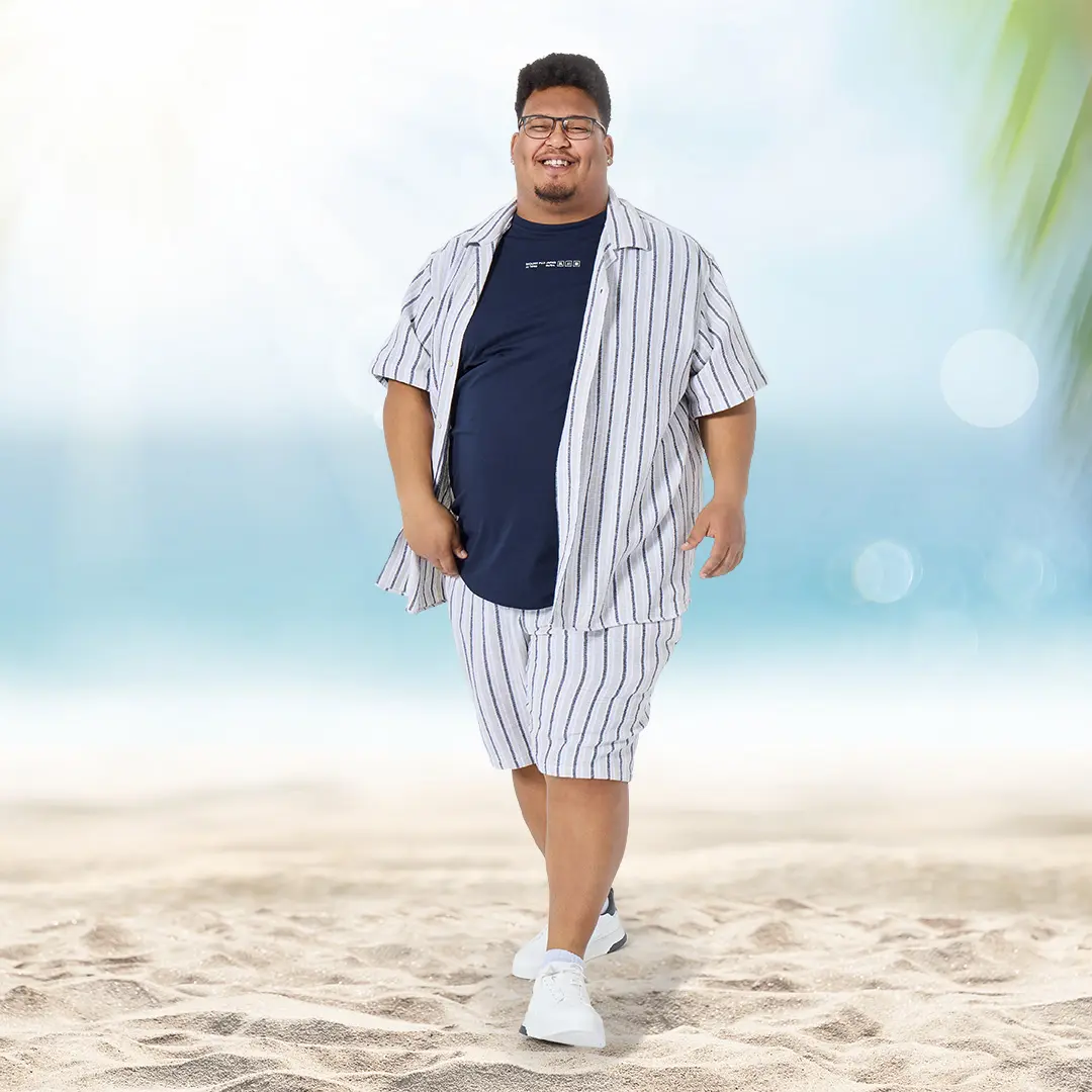Ein junger Mann mit Kurzarmhemd und Shorts im Streifenlook spaziert selbstbewusst am Strand – sommerliches Set für große Größen.