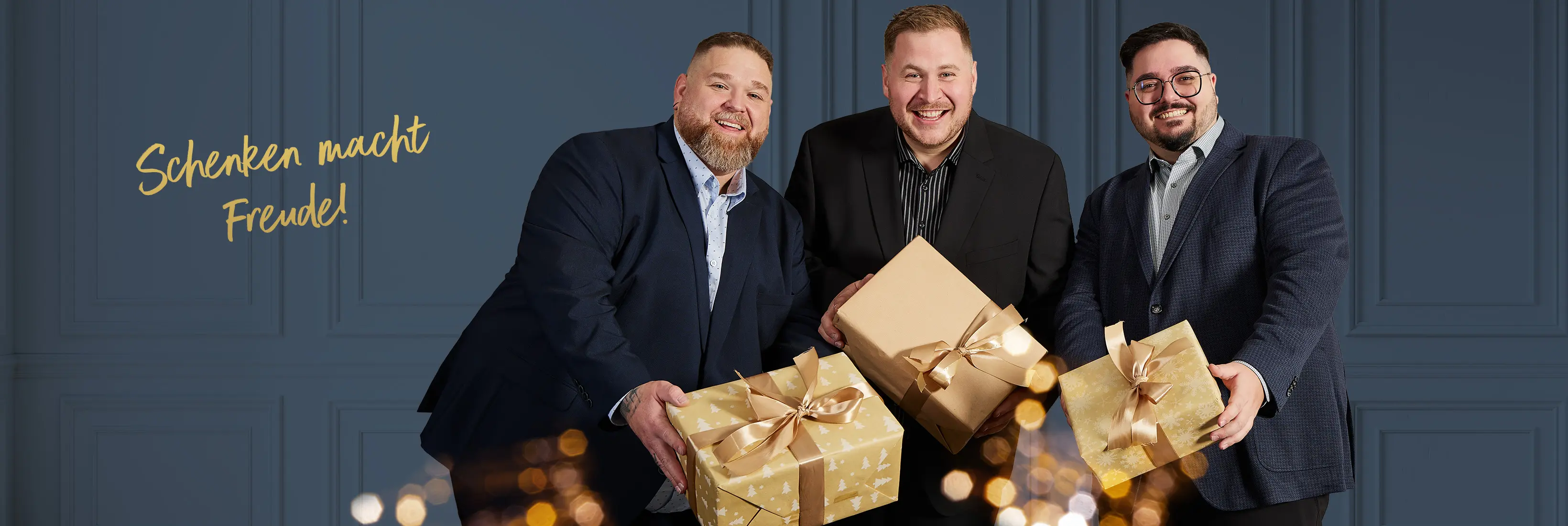 Drei Männer in eleganter Weihnachtsmode präsentieren verpackte Geschenke vor blauem Hintergrund.
