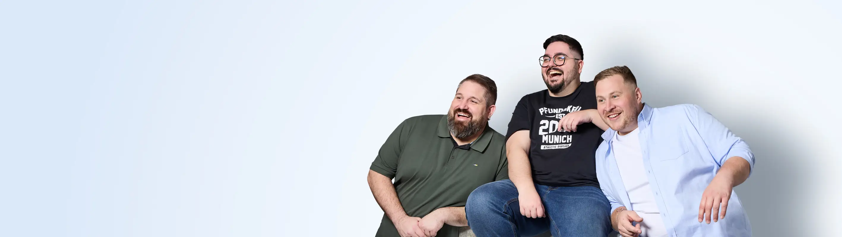 Drei gut gelaunte Männer in großen Größen sitzen gemeinsam vor hellem Hintergrund und tragen lässige Pfundskerl-Outfits