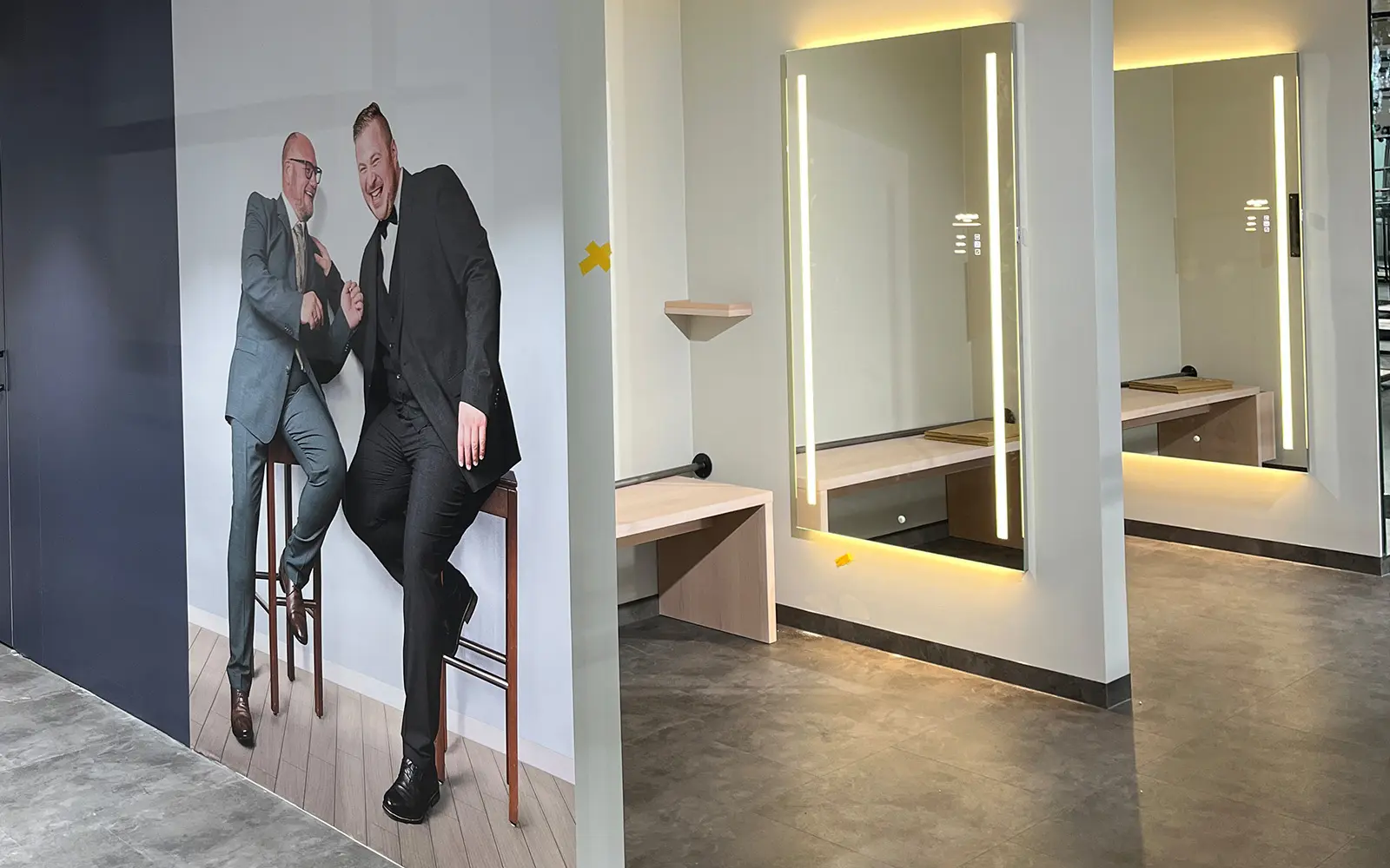 Modernisierter Umkleidebereich mit beleuchtetem Spiegel und großem Wandmotiv zweier Männer im Anzug