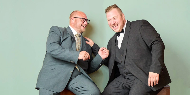 Zwei lachende Männer in eleganten Anzügen, einer in Grün mit gemusterter Krawatte, der andere in Dunkelgrau mit Fliege, gestikulieren vor grünem Hintergrund.