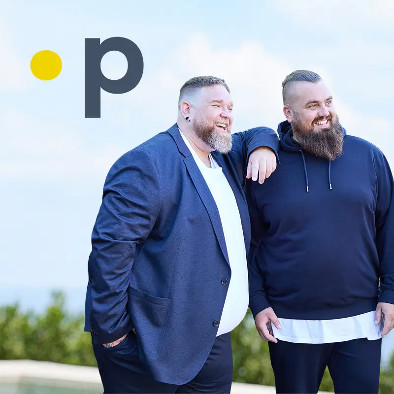 Zwei kräftige Männer lachen gemeinsam im Garten – einer im Sakko, einer im Hoodie