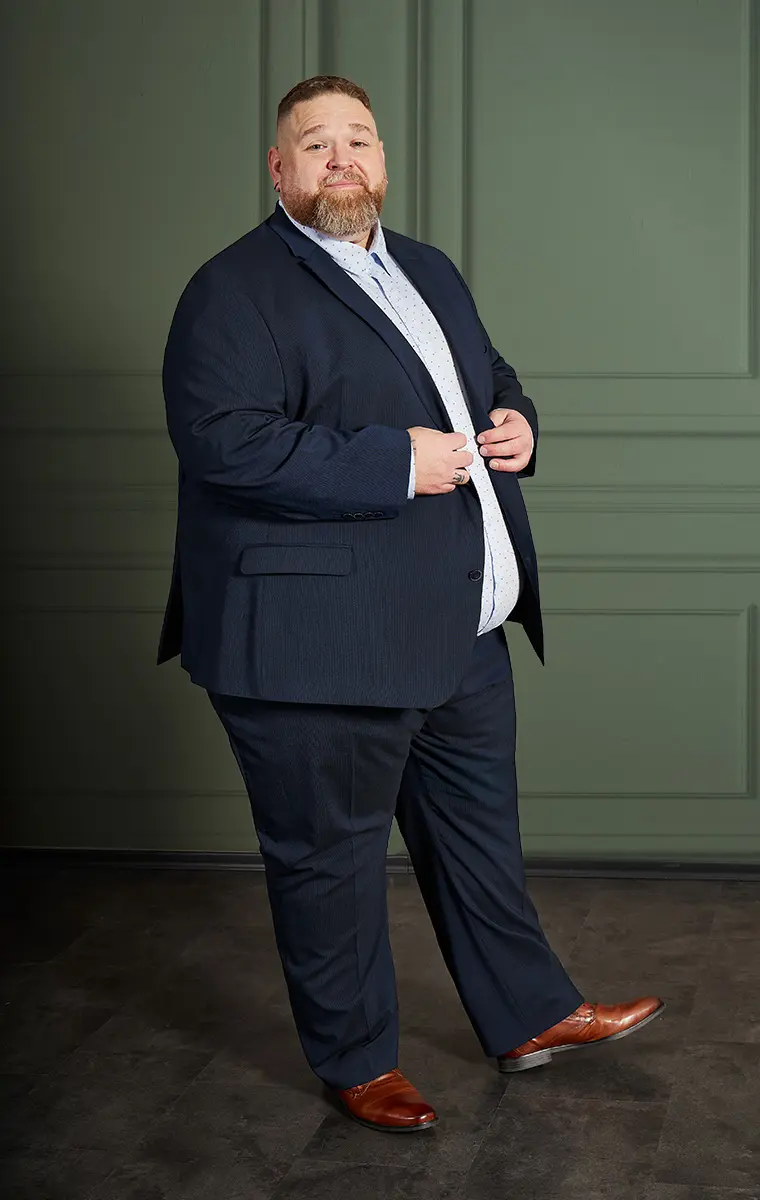 Mann in großer Größe im dunkelblauen Anzug mit braunen Schuhen vor grünem Hintergrund