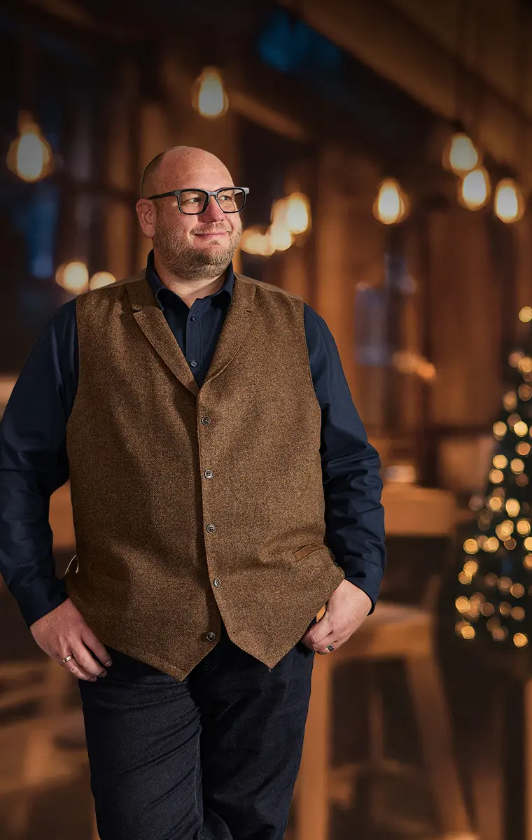 Mann in großer Größe mit brauner Wollweste und dunkelblauem Hemd in festlicher Umgebung