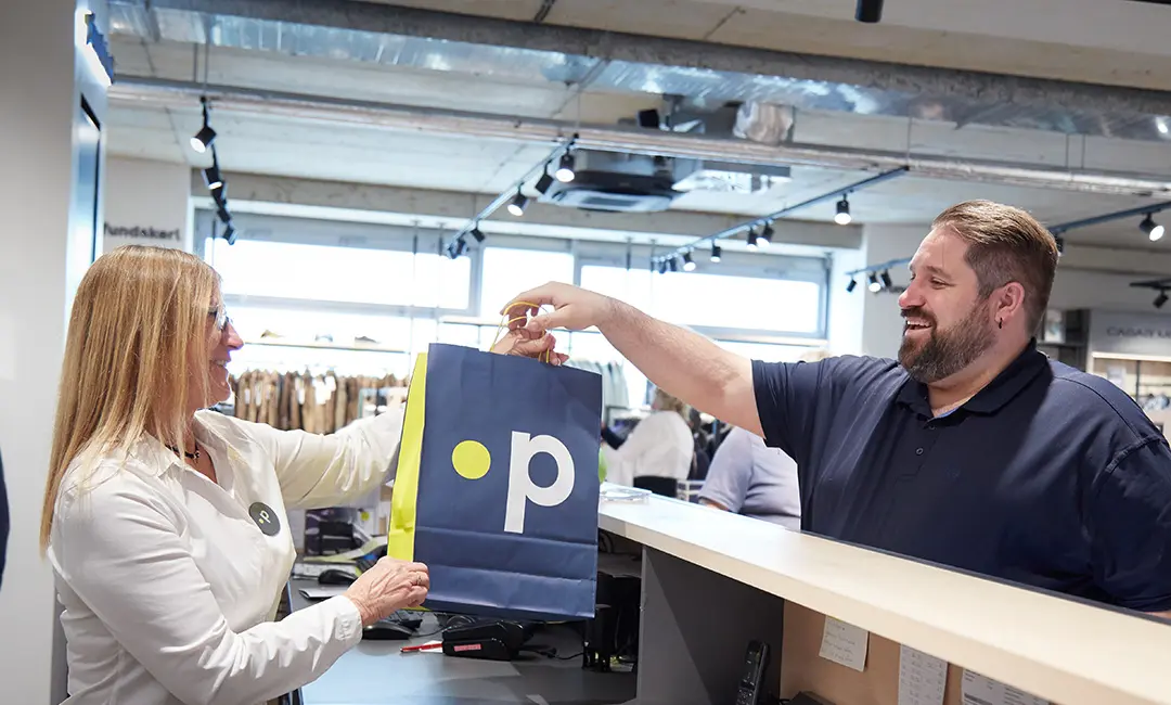 Frau überreicht in modernem Store einem Mann eine blaue Einkaufstasche mit Pfundskerl-Logo – im Hintergrund Kleidung und große Fenster.