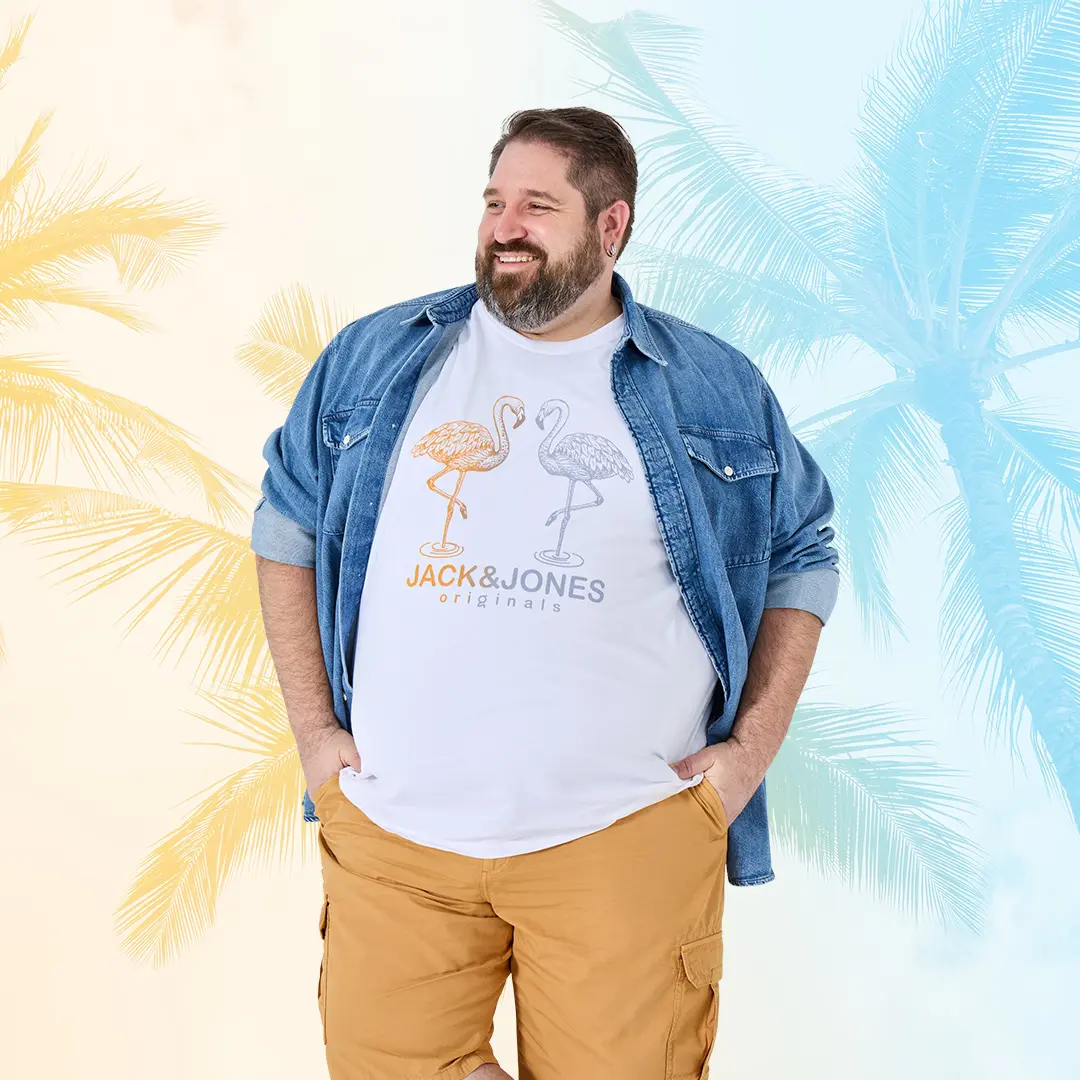 Mann mit lockerem Jeanshemd, weißem Printshirt und beigefarbenen Cargoshorts steht vor Palmen – sommerlich cool in großen Größen.