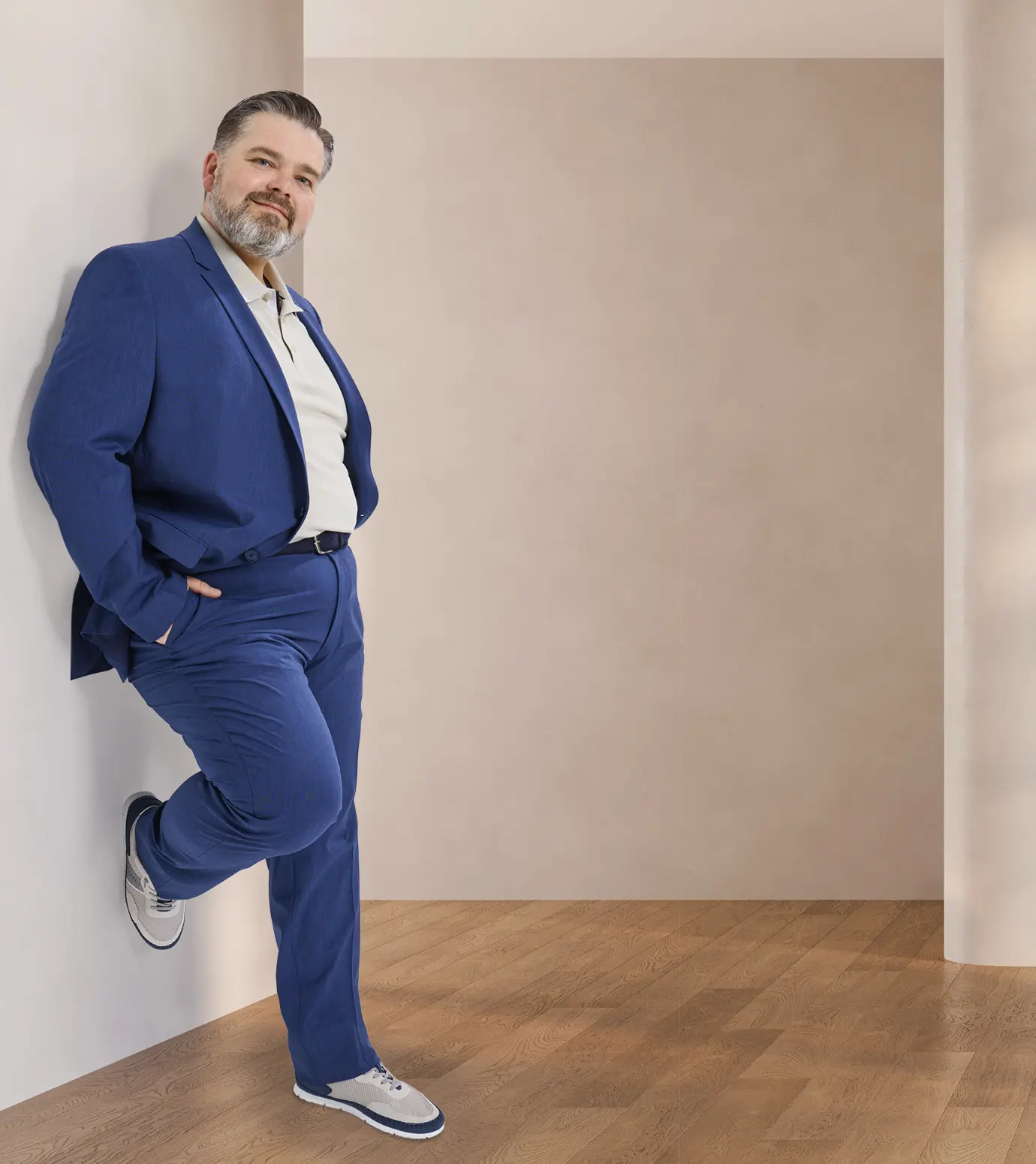 Person in blauem Anzug, weißem Hemd und weißen Sneakern mit blauen Details lehnt an Wand auf Holzfußboden.