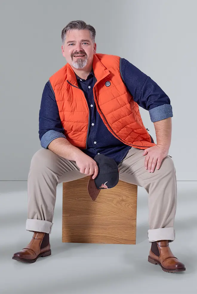 Model in großen Größen sitzt entspannt mit orangefarbener Steppweste, Jeanshemd und Chino